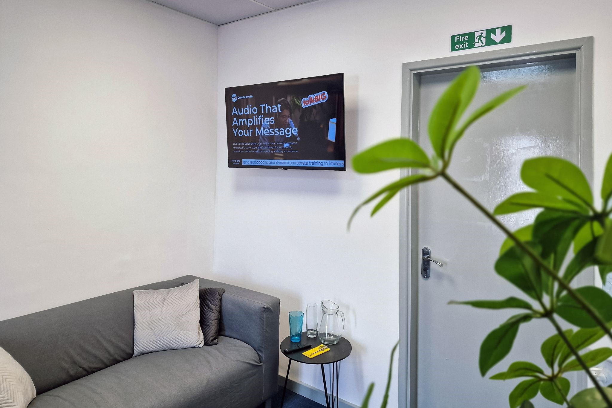 A modern office lobby features a gray sofa and a small round table with a glass pitcher and two glasses. A TV on the wall, powered by Yodeck, displays the text Audio That Amplifies Your Message. A plant partially obscures the view, adding a touch of greenery.