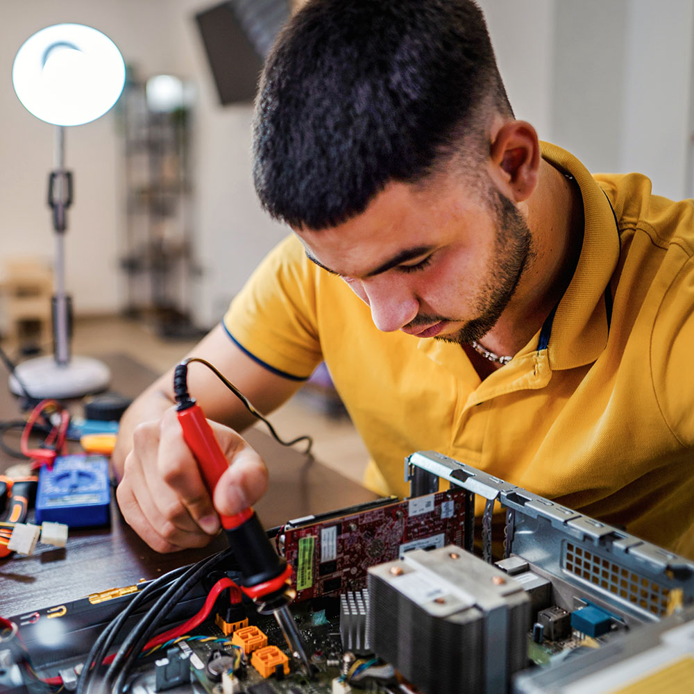 A young man in a yellow shirt works intently on a computer's internal components, holding a tool. A ring light illuminates his customer support workspace, which is scattered with electronic equipment and cables.